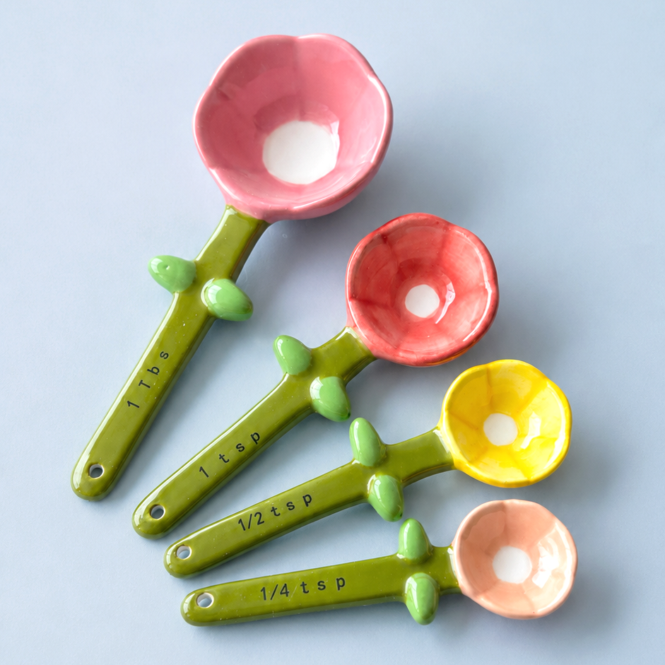 Four flower-shaped measuring spoons in pink, coral, yellow, and peach, arranged diagonally on a light blue background. The green handles have small leaf details and are printed with measurement markings.