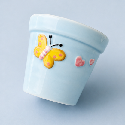 A light blue ceramic planter pot decorated with a yellow and pink butterfly and small pink hearts, shown on a solid light blue background.
