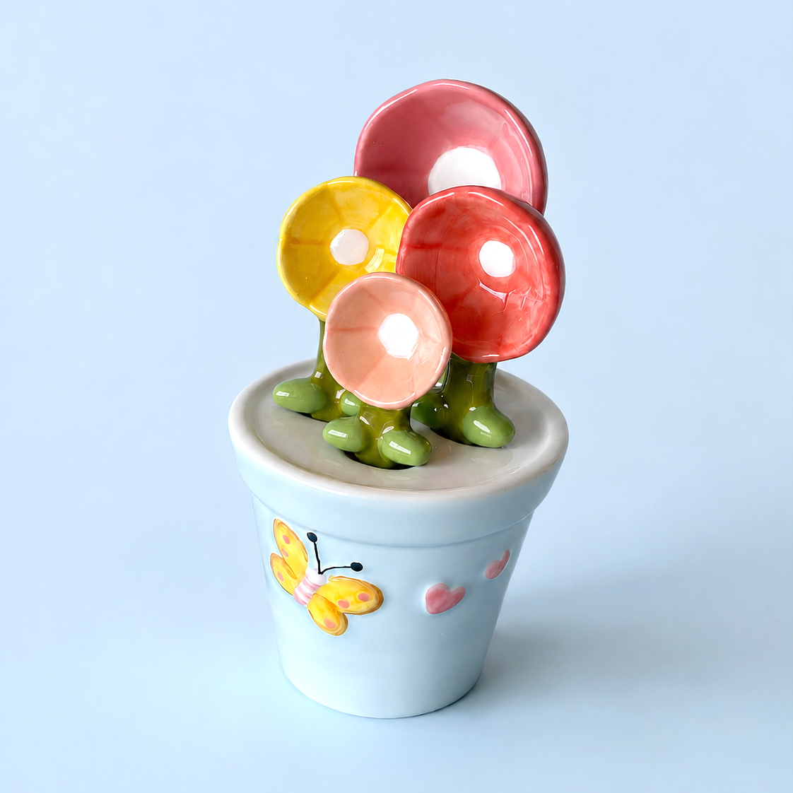 A ceramic flower pot figurine with three colorful flowers (pink, yellow, and coral red) on green stems, set against a solid light blue background.