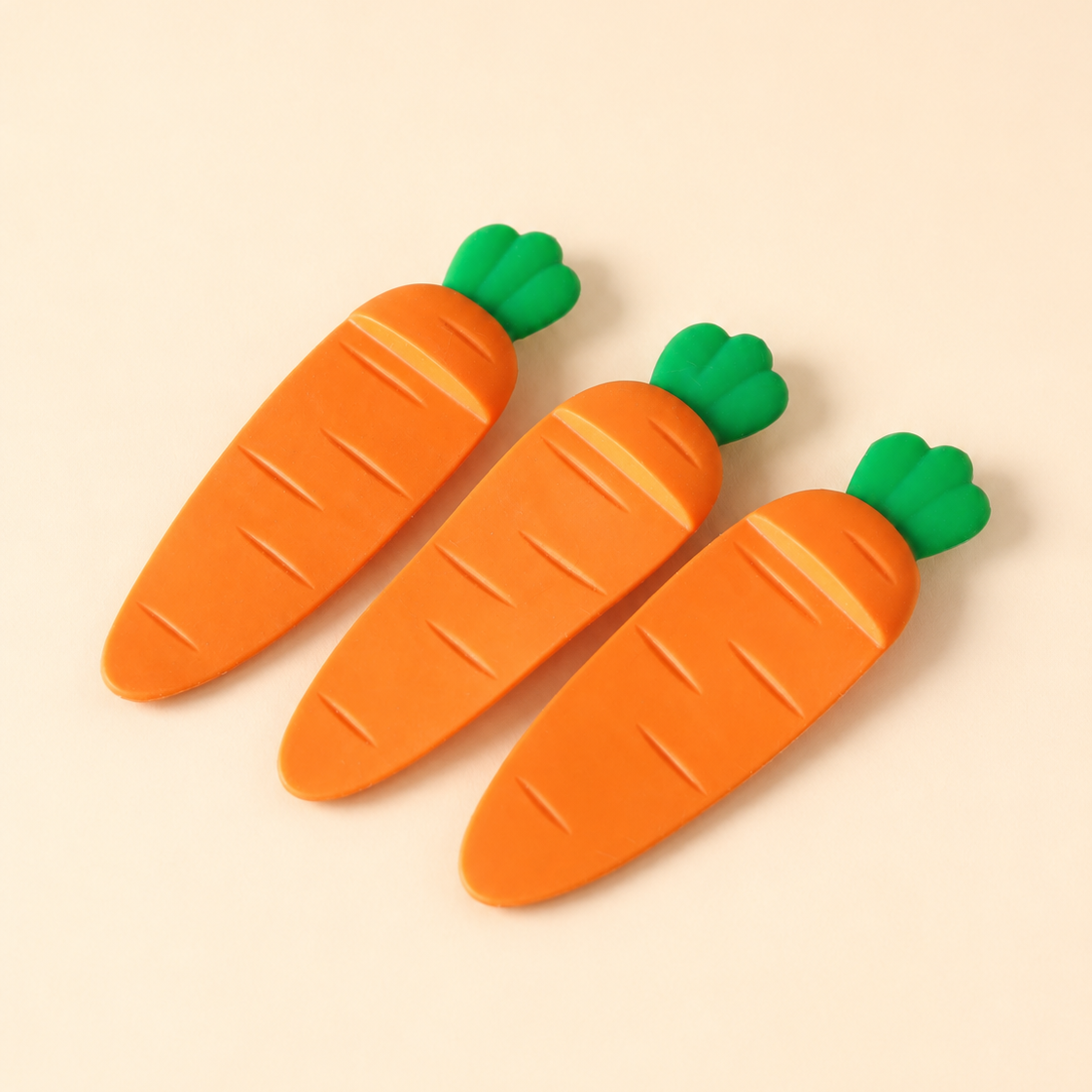 Three carrot-shaped carrot-shaped bookmarks with green tops on a wooden surface in orange and green, arranged diagonally on a solid beige background.