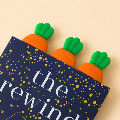 Three carrot-shaped bookmark with green tops and orange bodies peeking out from a navy blue book or box printed with gold stars and the partial text 'the rewind' on a warm beige background.
