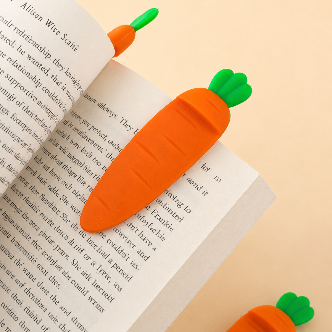 Two cute carrot-shaped bookmarks with green leafy tops resting on an open book page, set against a solid pale peach background.