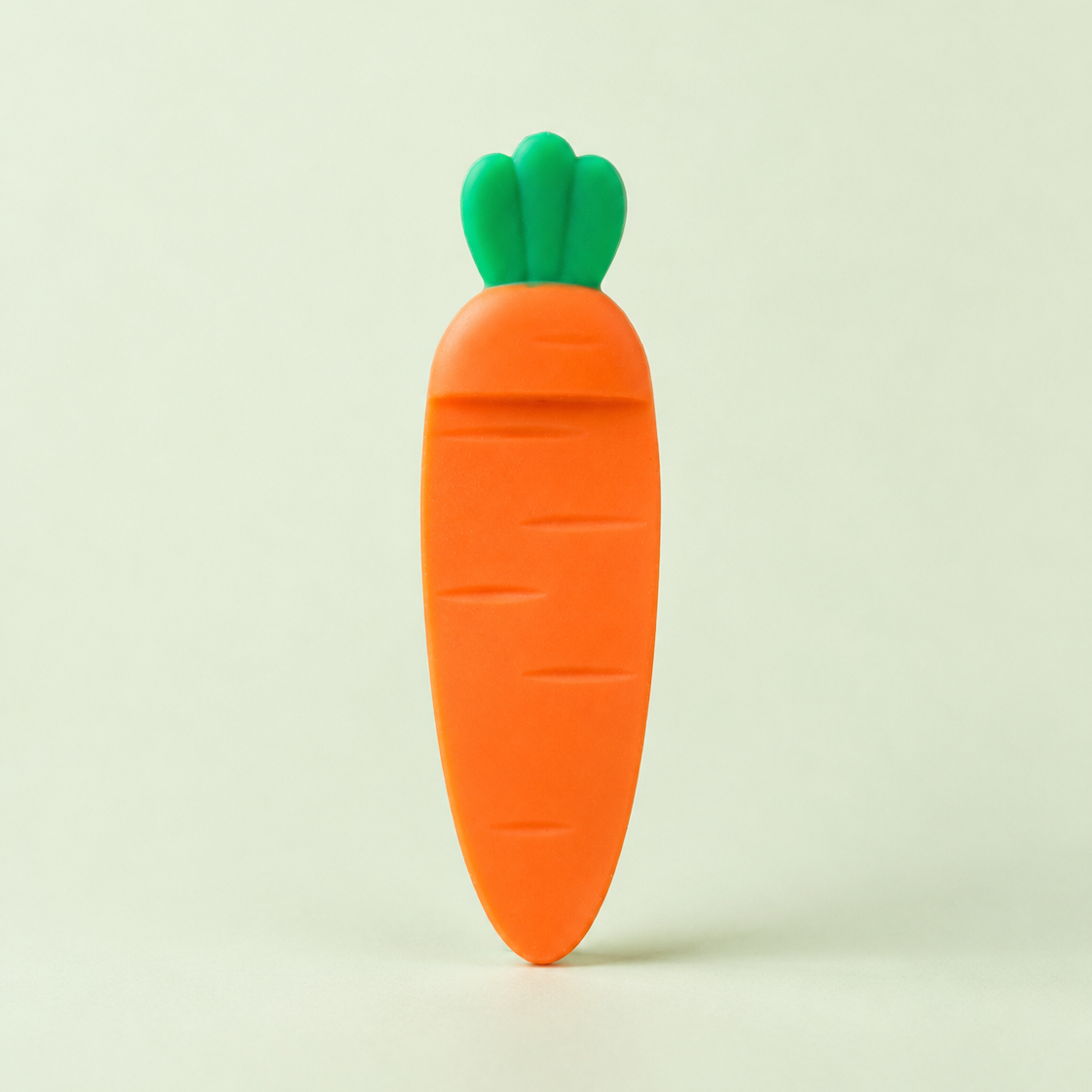 A small carrot-shaped carrot-shaped bookmarks with green tops on a wooden surface, standing upright on a light cream background. The carrot has a bright orange body with a green leafy top.