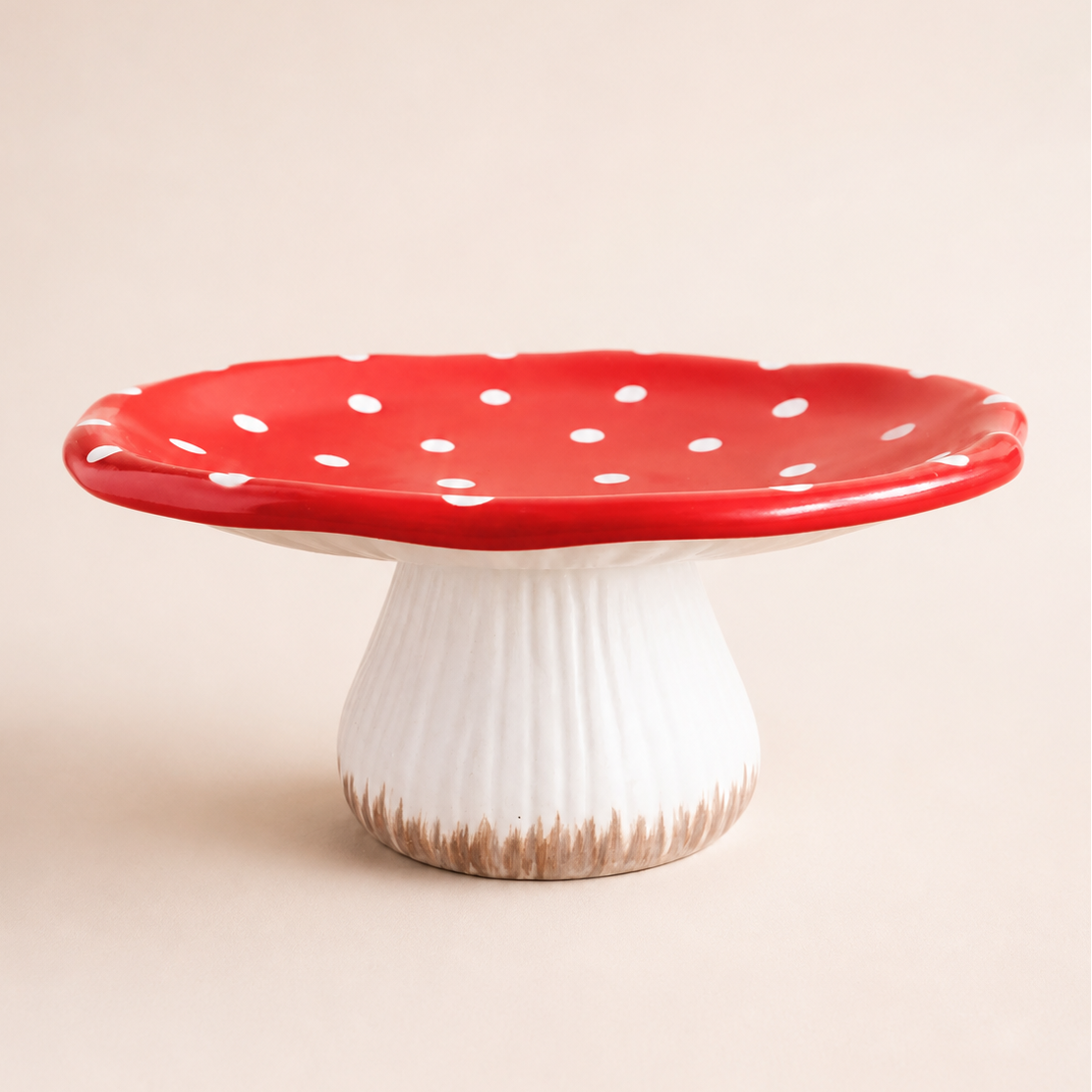 A mushroom-shaped ceramic cake stand with a glossy red cap dotted with white spots and a ribbed white stem, photographed on a plain beige studio background.
