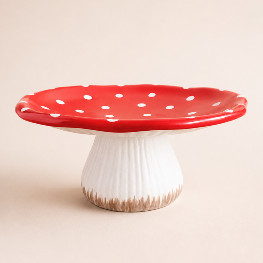 A mushroom-shaped ceramic cake stand with a glossy red cap dotted with white spots and a ribbed white stem, photographed on a plain beige studio background.