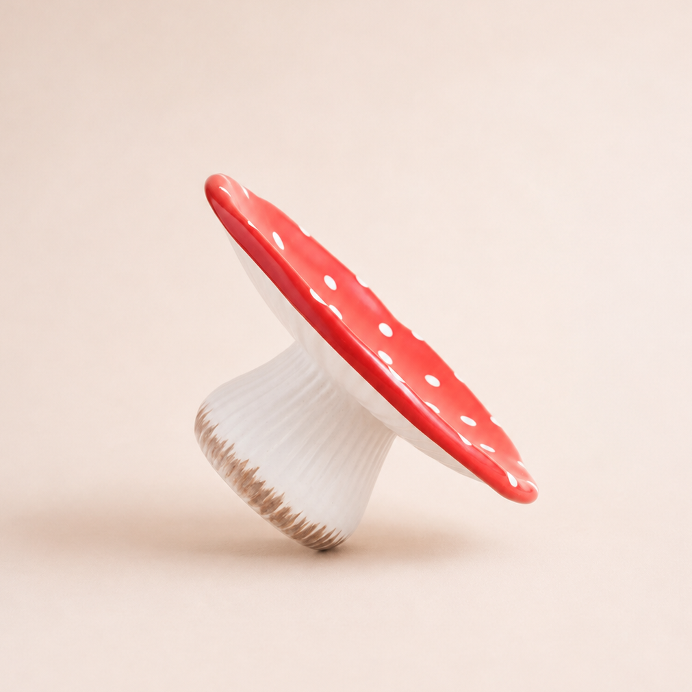 A small mushroom-shaped decorative product or cake stand with a glossy red cap and white polka dots, standing on a ribbed white base against a warm beige background.