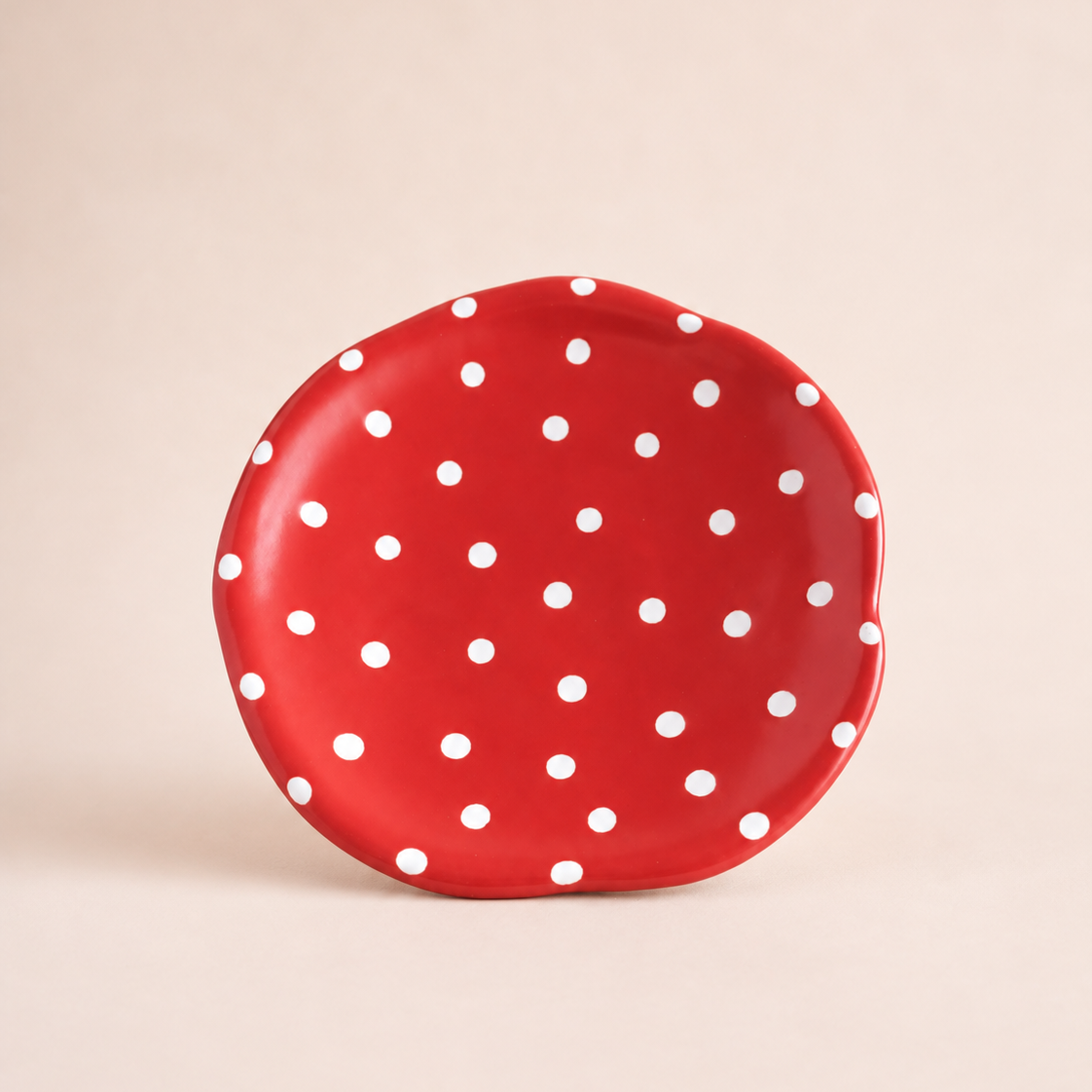 A small, scalloped-edge ceramic cake stand in vibrant red with a white polka dot pattern, set against a solid, warm beige background.