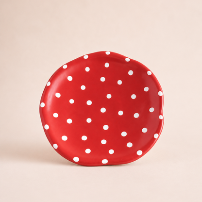 A small, scalloped-edge ceramic cake stand in vibrant red with a white polka dot pattern, set against a solid, warm beige background.