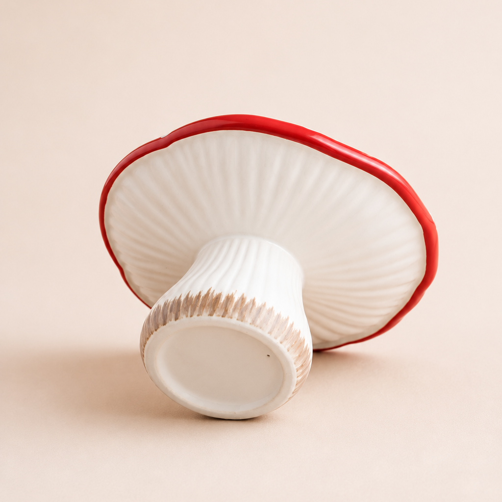 A mushroom-shaped ceramic cake stand with a ribbed white cap and stem, featuring a glossy red rim and a textured beige band near the base, set against a solid beige background.