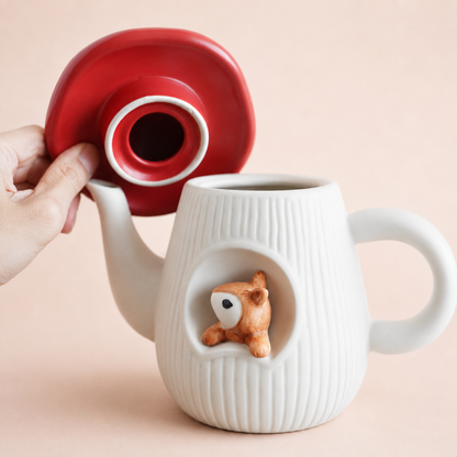 A white ribbed ceramic teapot, with a small brown squirrel peeking out from the front. A hand is lifting a red lid to show the mug's interior, against a soft beige background.