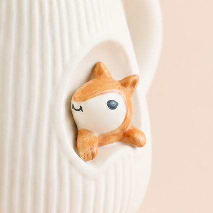 A white ribbed ceramic teapot with a small orange and white cartoon squirrel peeking out from the side, set against a solid pale pink background.