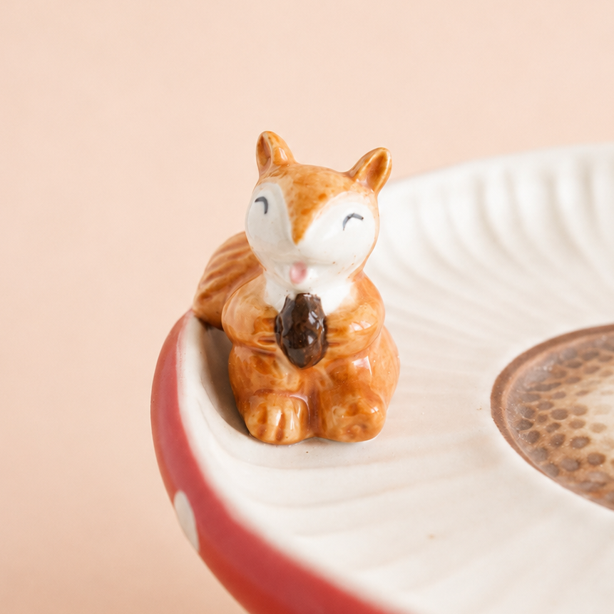A small ceramic squirrel figurine holding an acorn, displayed on a red and white mushroom-shaped dish against a soft beige background.