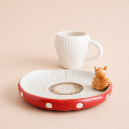 A ceramic coffee table tray with a red rim, white polka dots, and a textured off-white center, styled with a small white ribbed mug and a miniature orange squirrel figurine on a warm beige background.