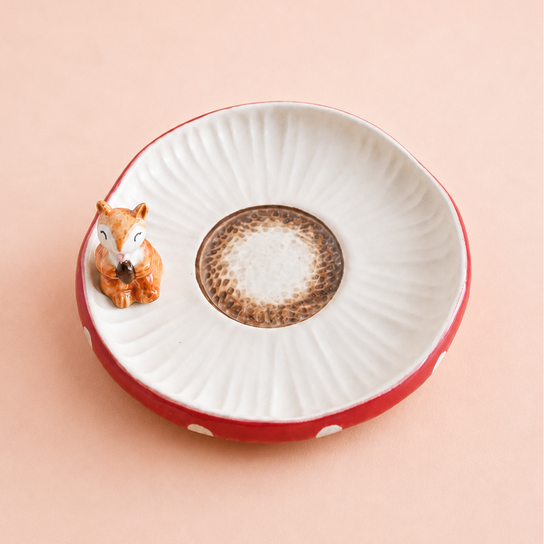A small ceramic dish with a white ribbed interior, a red exterior, a brown speckled center, and a small squirrel figurine sitting on the left edge, set against a solid pale peach background.