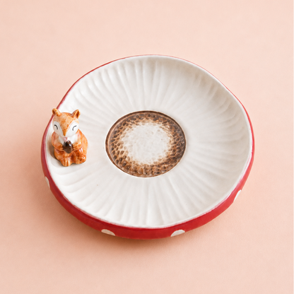 A small ceramic dish with a white ribbed interior, a red exterior, a brown speckled center, and a small squirrel figurine sitting on the left edge, set against a solid pale peach background.