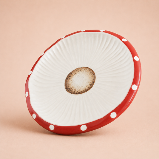 A round ceramic plate with a white ribbed interior, a brown textured center, and a red exterior rim decorated with white polka dots, set against a solid beige background.