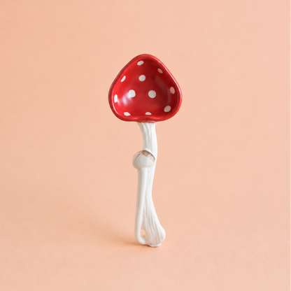 A ceramic mushroom-shaped spoon with a glossy red cap decorated with white polka dots and a textured white stem, set against a solid peach-pink background.