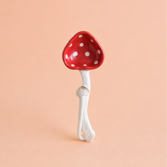 A ceramic mushroom-shaped spoon with a glossy red cap decorated with white polka dots and a textured white stem, set against a solid peach-pink background.