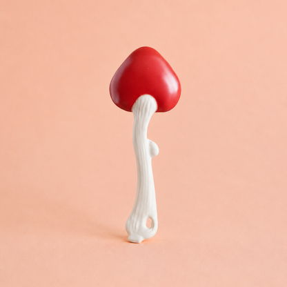 A mushroom-shaped spoon with a glossy red cap and a textured white stem, standing upright against a solid peach-pink background.
