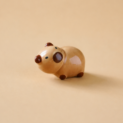 A small ceramic animal figurine, shaped like a capybara, in a glossy light beige glaze with dark brown accents on its snout, ears, eyes, and feet, set against a solid warm beige background.