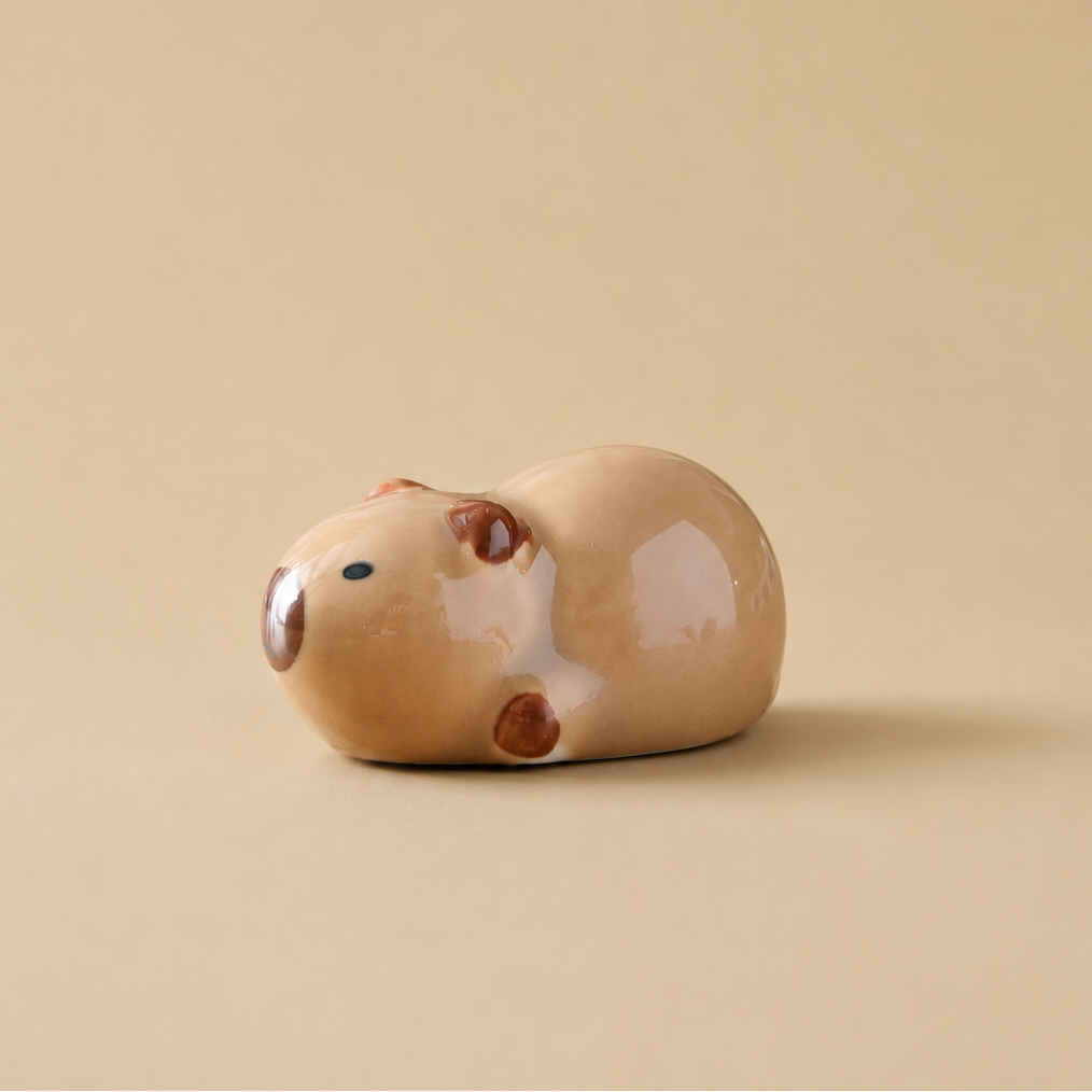 A small glossy ceramic animal figurine, shaped like a capybara, in a light tan glaze with darker brown accents, set against a solid beige background.
