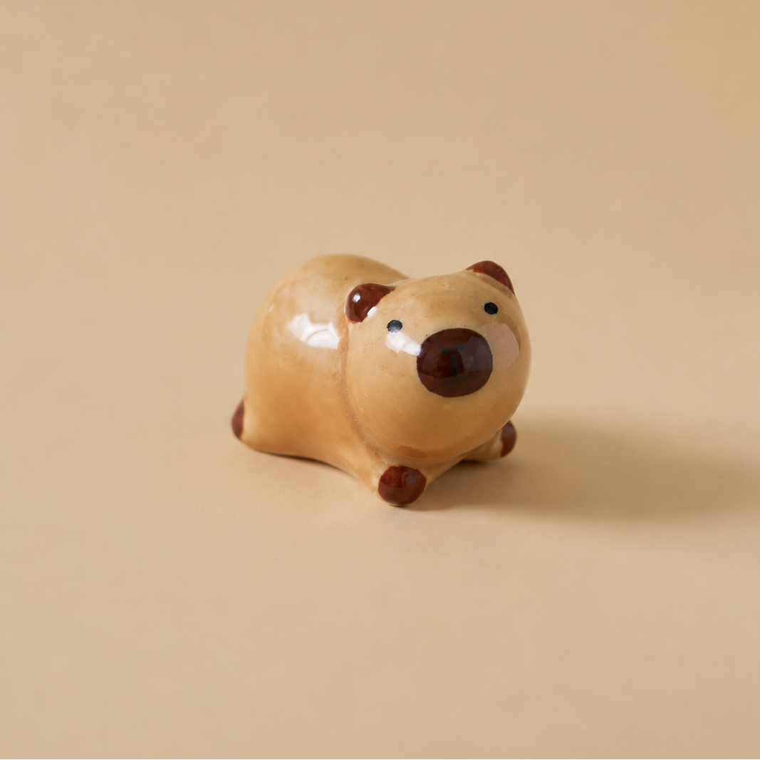 A small ceramic animal figurine, shaped like a seated capybara, in a glossy light tan glaze with dark brown accents on its snout, paws, and ears, set against a solid warm beige background.