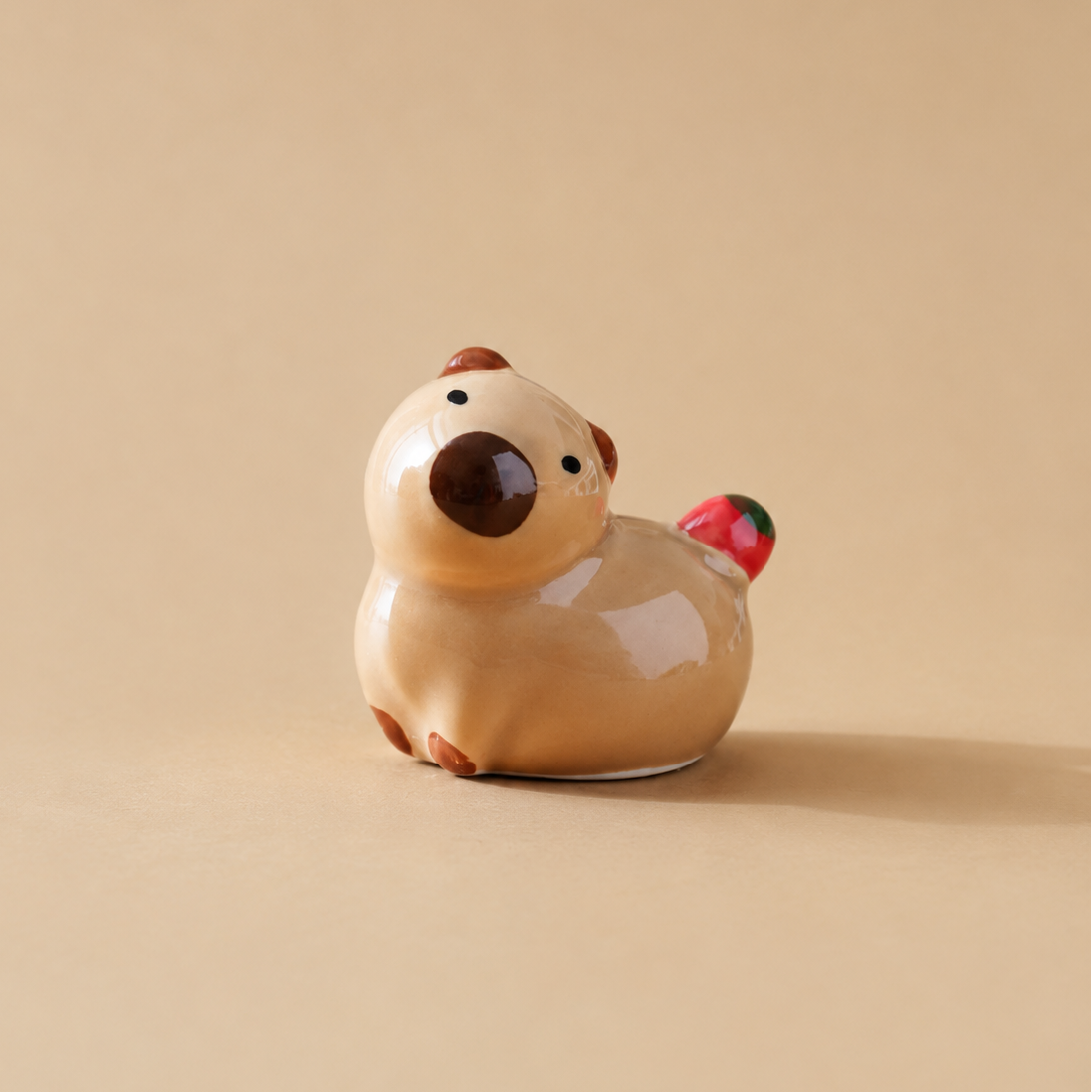 A small glossy ceramic figurine of a cartoon animal capybara with a beige body, dark brown snout and ears, and a tiny red-and-black accent on its tail, photographed on a warm beige background.