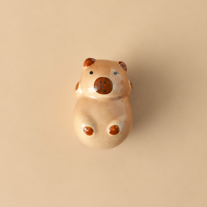 A small ceramic figurine of a capybara with a glossy finish, featuring a light beige body with darker brown spots and a dark brown snout, centered on a solid beige background.