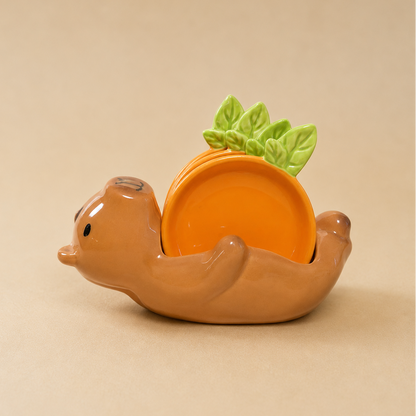 A ceramic capybara-shaped dish holder in warm brown, holding a small orange plate with green leaf accents, set against a solid beige background.