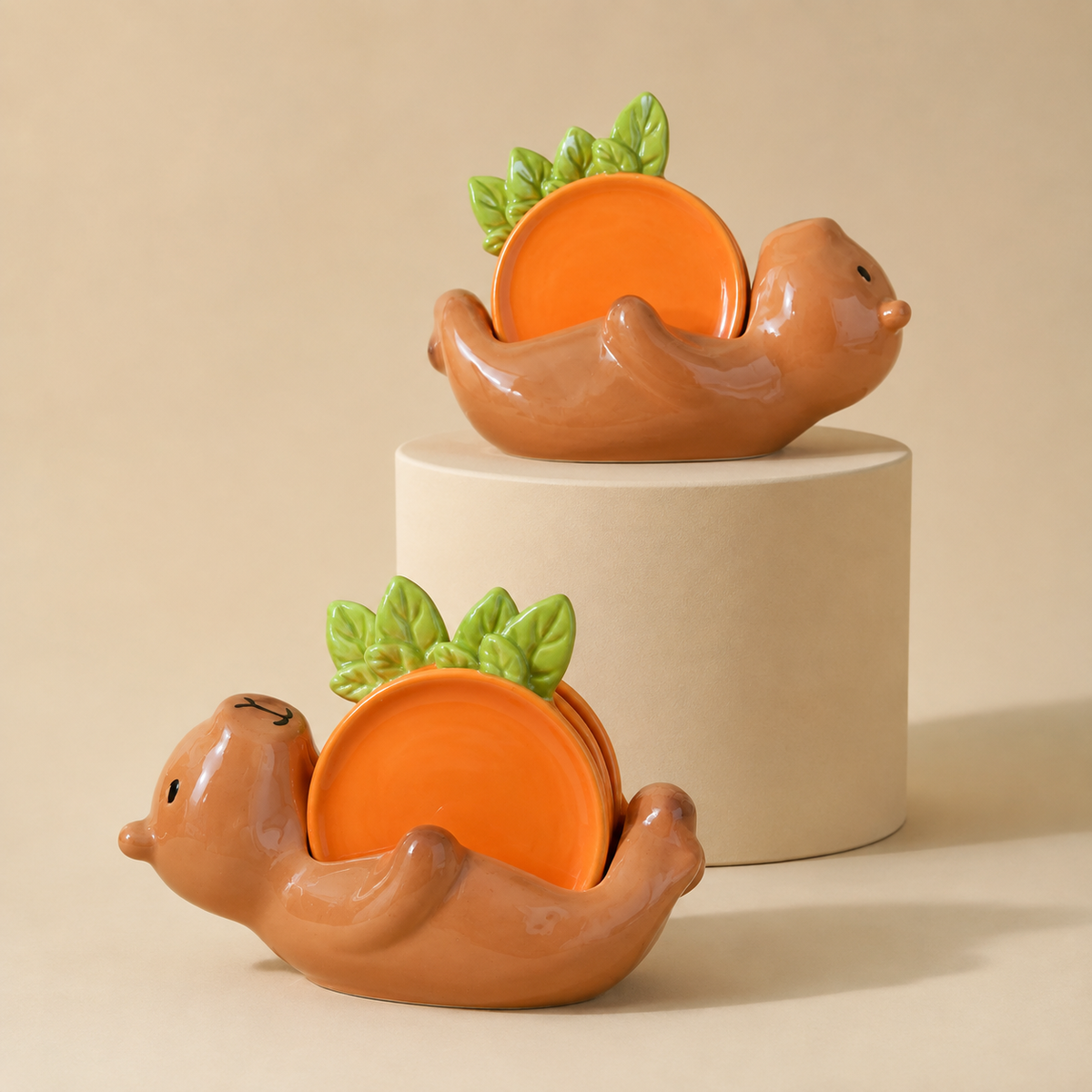 Two small ceramic capybara-shaped dish holders in a warm terracotta brown, each holding a round orange plate with green leaf accents, displayed on a beige pedestal against a beige background.