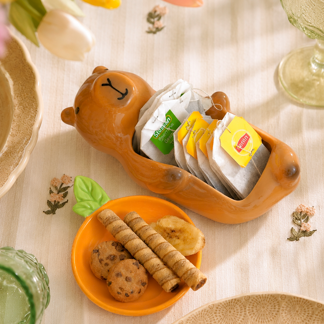 A ceramic capybara-shaped tea bag holder filled with assorted L'Englez tea bags, styled on a table with cookies on a small orange plate and flowers in the background.