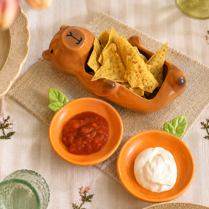 A styled tabletop scene with a ceramic capybara-shaped tortilla chip dish holding yellow chips, alongside two small orange bowls containing red salsa and white sour cream on a beige tablecloth with flowers and glassware in the background.