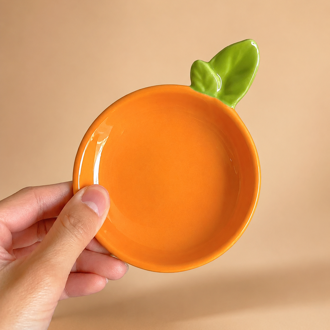 An orange-shaped ceramic dish with a green leaf handle, held by a hand against a solid beige background.