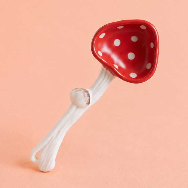 A small mushroom-shaped spoon with a glossy red cap covered in white polka dots and a white stem, photographed on a solid peach-pink background.