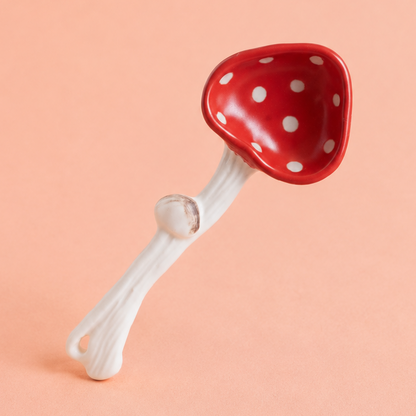 A small mushroom-shaped spoon with a glossy red cap covered in white polka dots and a white stem, photographed on a solid peach-pink background.