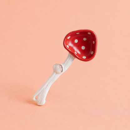 A small mushroom-shaped spoon with a glossy red cap covered in white polka dots and a white stem, photographed on a solid peach-pink background.