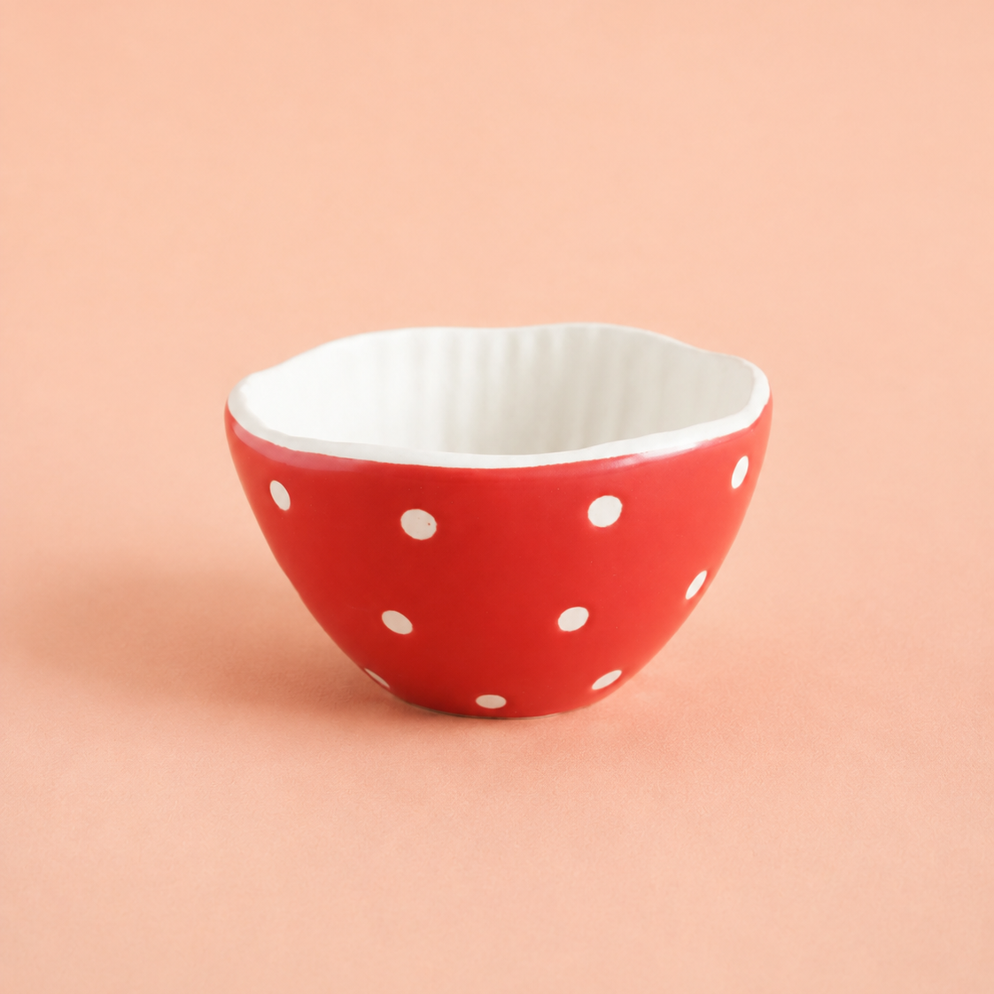 A small ceramic bowl with a red exterior decorated with white polka dots and a white ribbed interior, set against a solid peach-pink background.