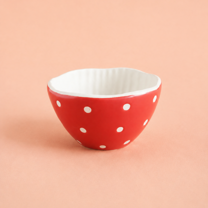 A small ceramic bowl with a red exterior decorated with white polka dots and a white ribbed interior, set against a solid peach-pink background.