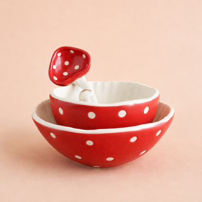 A set of two stacked ceramic bowls and a matching spoon in red with white polka dots, set against a solid pale peach background. Mushroom theme tableware.