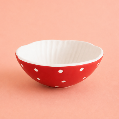 A small ceramic bowl with a scalloped rim, featuring a red exterior with white polka dots and a white ribbed interior, set against a solid peach-pink background.