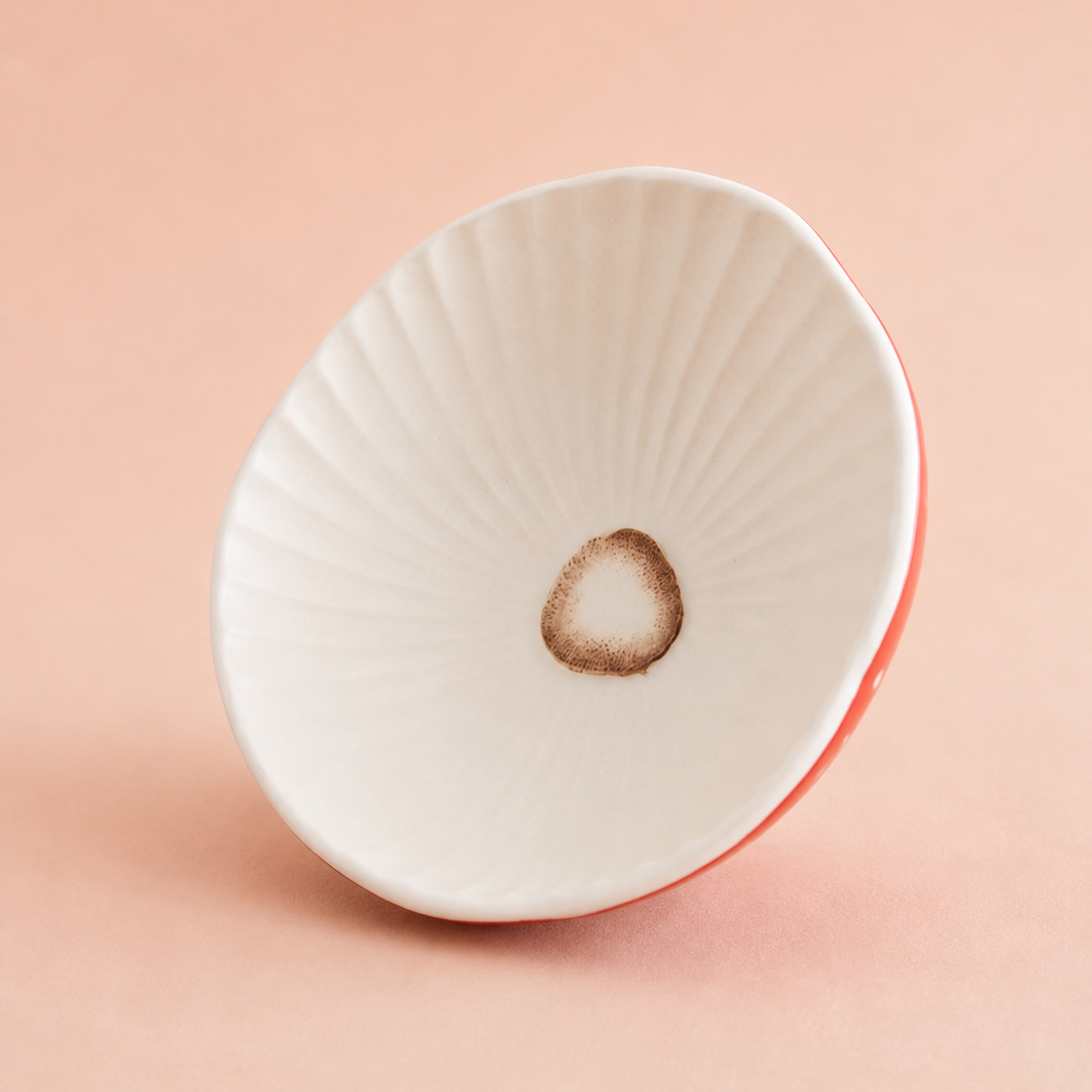 A white ceramic bowl with a delicate radial rib pattern and a small brown circular mark in the center, shown on a solid peach-pink background.