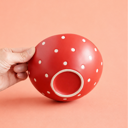 A hand holding a glossy red ceramic bowl with white polka dots, set against a solid peach-colored background.