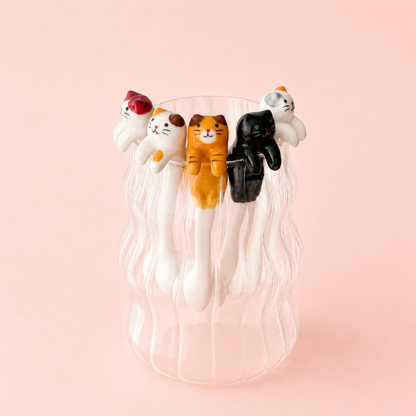 A set of five cat-shaped spoons in various colors (white, orange, black, and calico) arranged on the rim of a clear ribbed glass cup against a solid pale pink background.