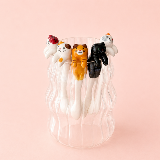 A set of five cat-shaped spoons in various colors (white, orange, black, and calico) arranged on the rim of a clear ribbed glass cup against a solid pale pink background.