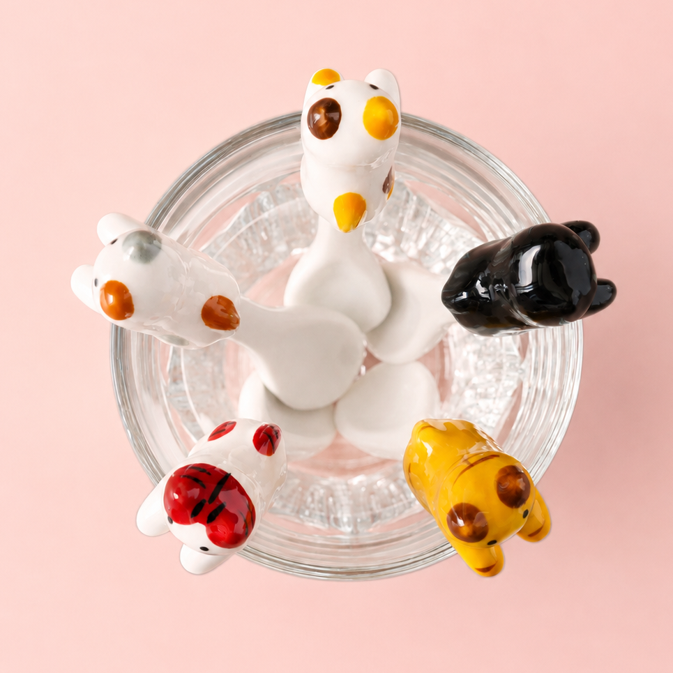 Four small animal cat figurines and teaspoons arranged around a clear glass bowl on a soft pink background. The figurines resemble stylized cats or small mammals in white, black, yellow, and red tones with orange and yellow facial details.
