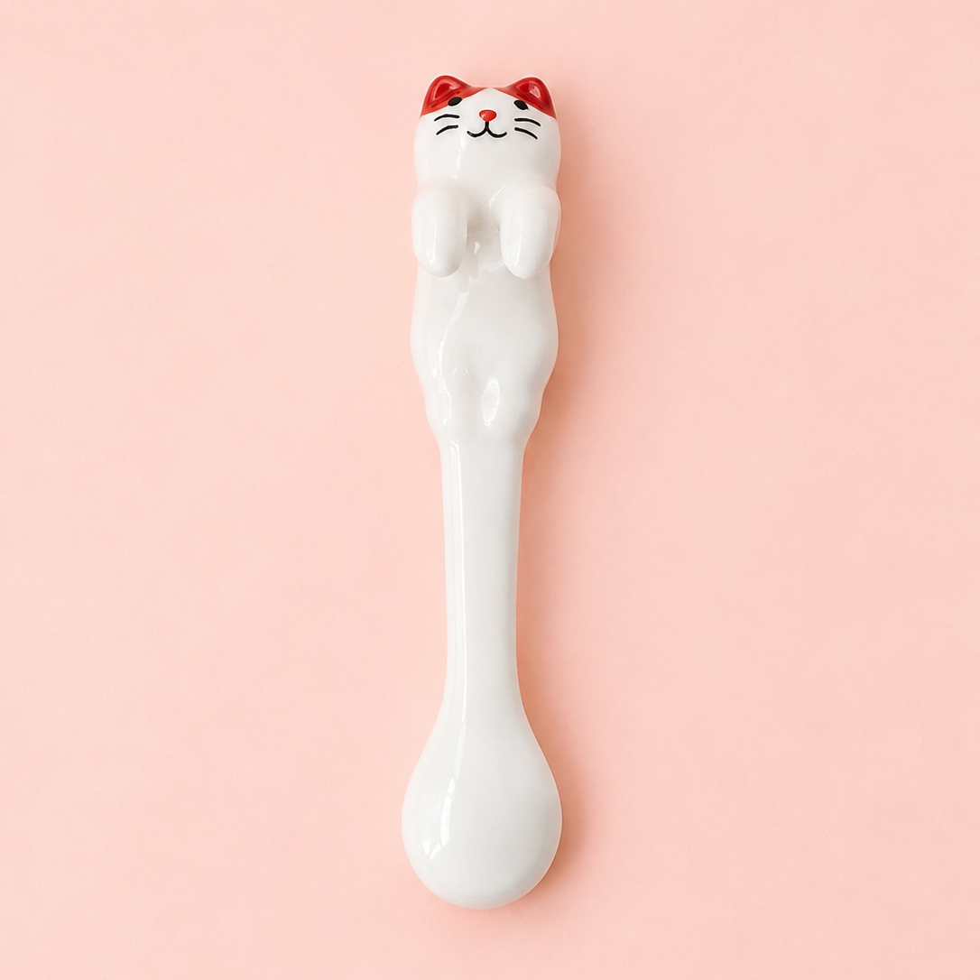 A white ceramic spoon with a cute cat figurine on the handle, featuring red ears and a black face, set against a solid pink background.