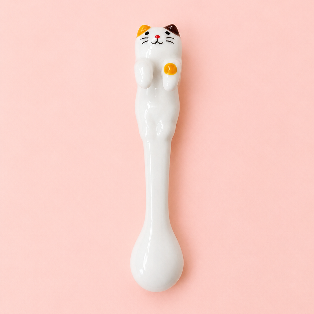 A white ceramic spoon shaped like a cat, featuring a detailed face with orange, black, and white markings and a raised yellow paw, set against a solid light pink background.