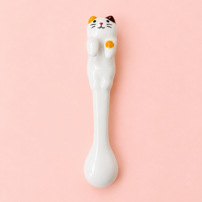A white ceramic spoon shaped like a cat, featuring a detailed face with orange, black, and white markings and a raised yellow paw, set against a solid light pink background.