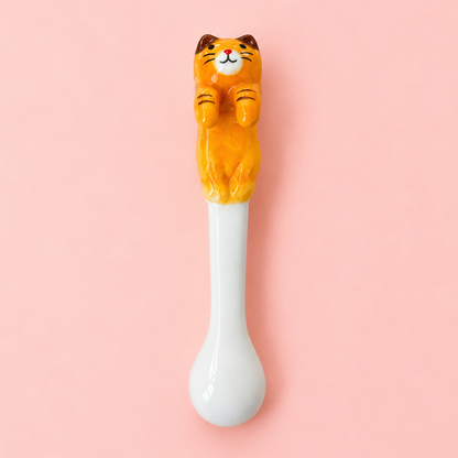 A ceramic spoon with a glossy white bowl and a small orange tabby cat figurine perched on the spoon handle, set against a solid pink background.