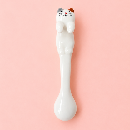 A white ceramic spoon with a cute cat figurine on the handle, featuring a white body with orange and gray ears and a black whisker face, set against a solid pink background.